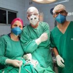 A imagem mostra parte da equipe responsável pela cirurgia, Nela há três pessoas com uniforme verde, touca, máscara e luva.