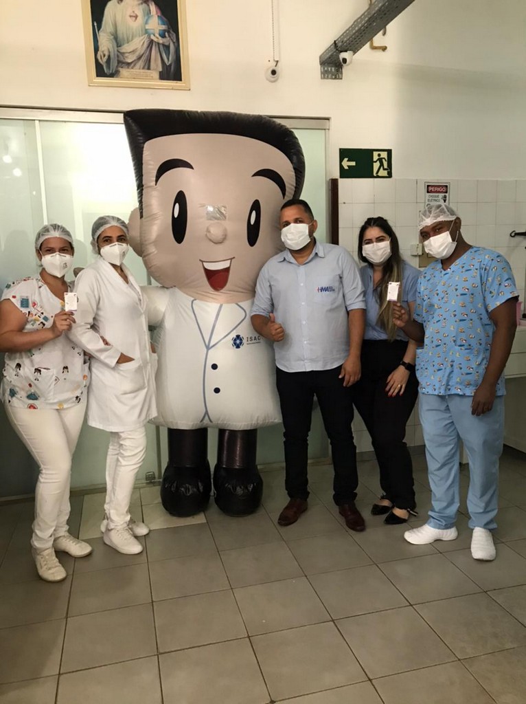 Pessoas tiram foto ao lado do Isaquinho, boneco que representa a marca ISAC. Todos estão com máscara, touca e fardamento hospitalar.
