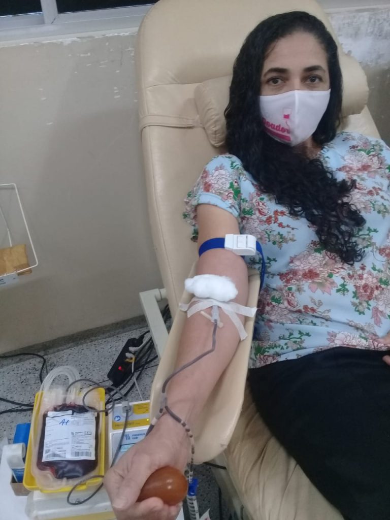 Mulher sentada numa poltrona hospitalar doando sangue. Ela usa máscara e sorri para câmera.