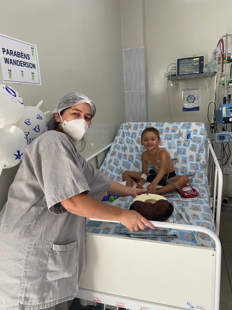 Mulher com fardamento hospitalar na cor cinza está ao lado de um leito. Deitado no leito, aparece uma criança que sorri para foto.