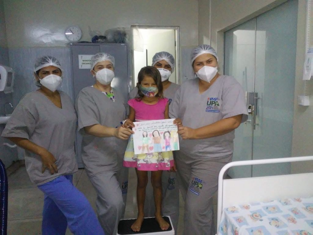 quatro colaboradores do HMA posam para foto ao lado da pequena paciente. Todos usam máscara .
