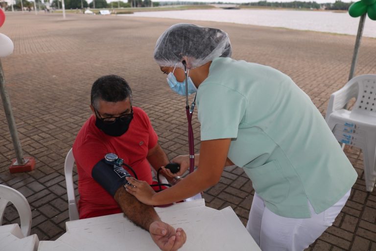 Um homem de camisa vermelha está sentado enquanto uma mulher de frente para ele faz a aferição da pressão arterial com o aparelho no braço direito do homem.